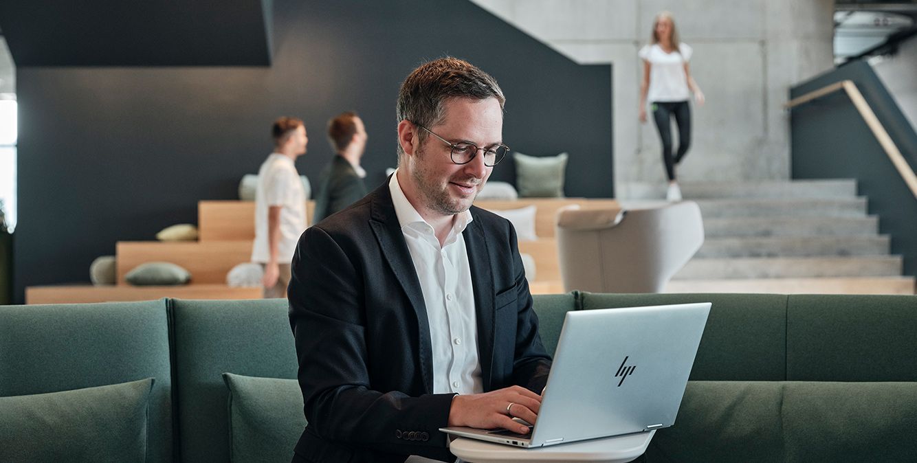 Mitarbeiter von Streit Service & Solution sitzt in der Arbeitswelt und arbeitet am Laptop. Hinter ihm laufen weiter Mitarbeitende.