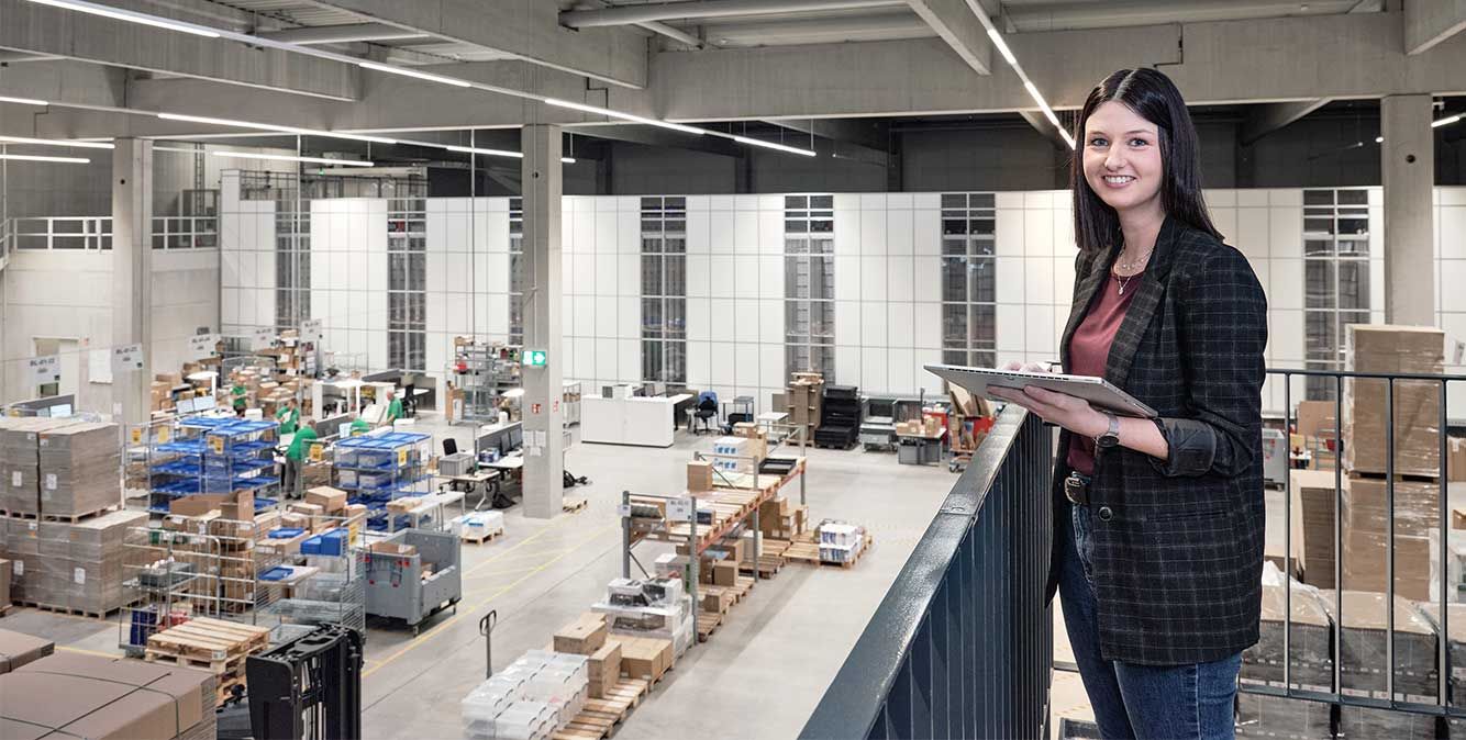 Mitarbeiterin von Streit Service & Solution steht im modernen Logistikzentrum und arbeitet an einem Tablet. Im Hintergrund ist die Autostore Anlage zu sehen.