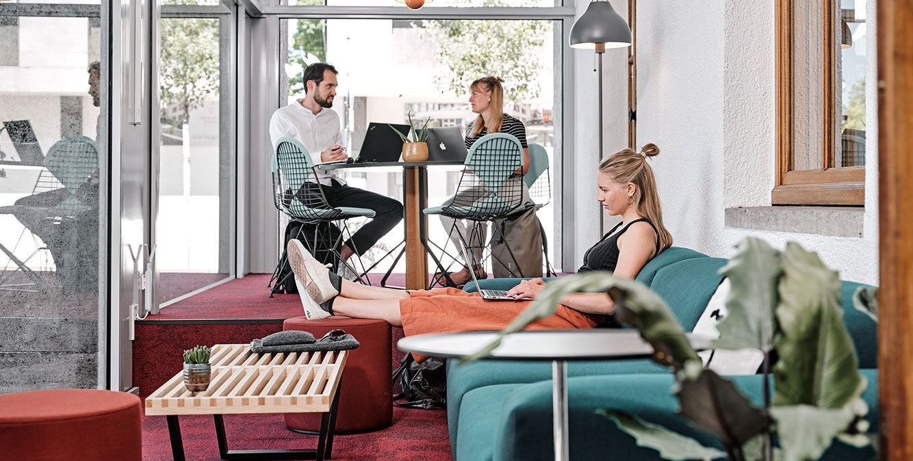 Eine Frau sitzt auf einem Sofa und arbeitet am Laptop. Im Hintergrund sitzen zwei weitere Personen zusammen an einem Tisch und arbeiten am Laptop.