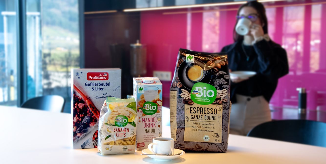 Produkte von dm stehen auf einem Tisch. Im Hintergrund trinkt eine Frau Kaffee.