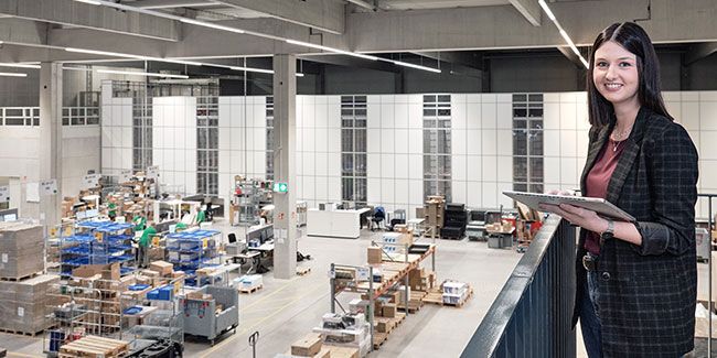 Mitarbeiterin von Streit Service & Solution steht im modernen Logistikzentrum und arbeitet an einem Tablet. Im Hintergrund ist die Autostore Anlage zu sehen.