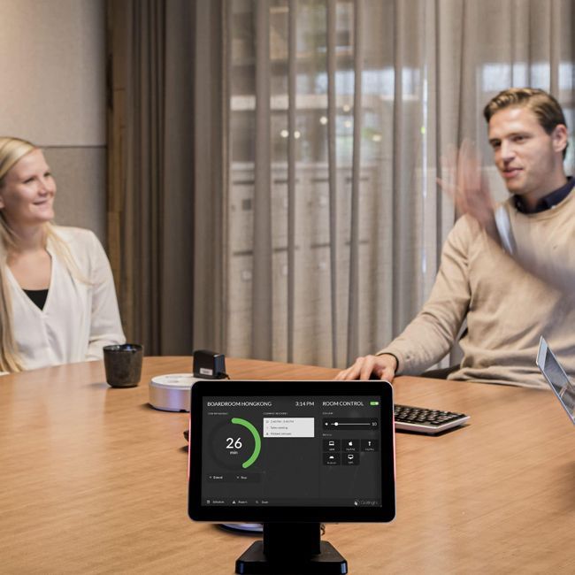 Personen sitzen an einem Besprechungstisch auf dem ein Room Panel steht