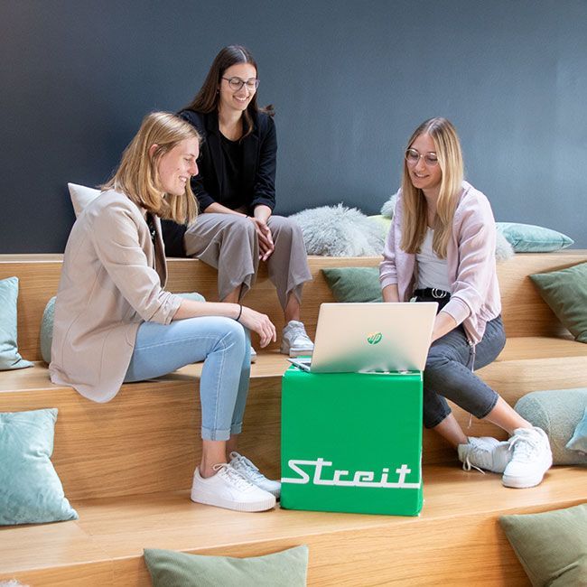 Duale Studentinnen von Streit Service & Solution sitzen auf einer Treppe und schauen zusammen auf einen Laptop