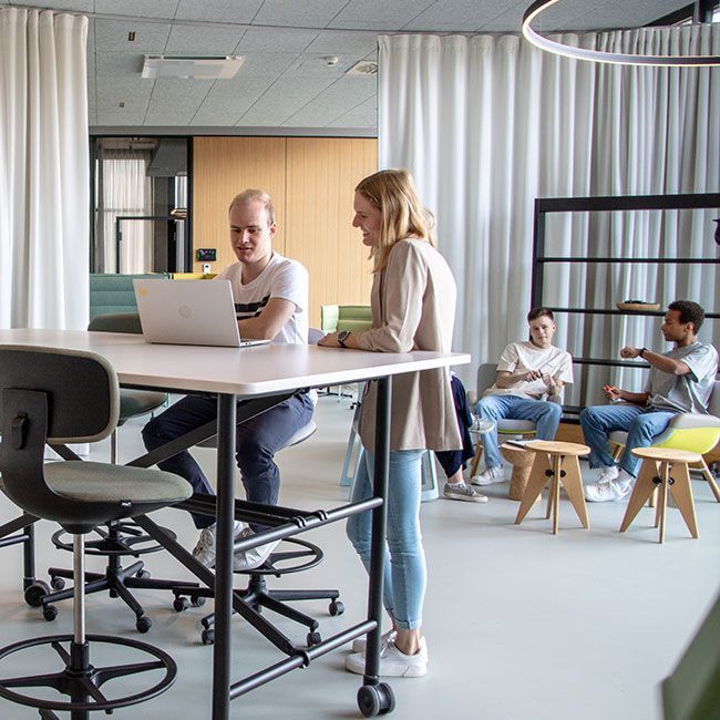Duale Studenten von Streit Service & Solution arbeiten zusammen. Zwei stehen an einem Tisch und schauen zusammen in einen Laptop. Im Hintergrund sitzen drei weitere Studenten zusammen und sprechen miteiander