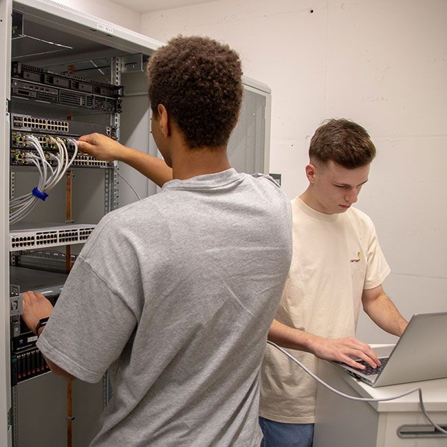 Zwei Auszubildende von Streit Service & Solution arbeiten zusammen im Serverraum. Einer arbeitet direkt am Server während der andere am Laptop schreibt