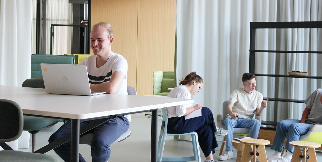 Auszubildende von Streit Service & Solution arbeiten zusammen in der Arbeitswelt. Einer sitzt an einem Tisch und arbeitet am Laptop. Die anderen sitzen im Hintergrund und sprechen miteinander.