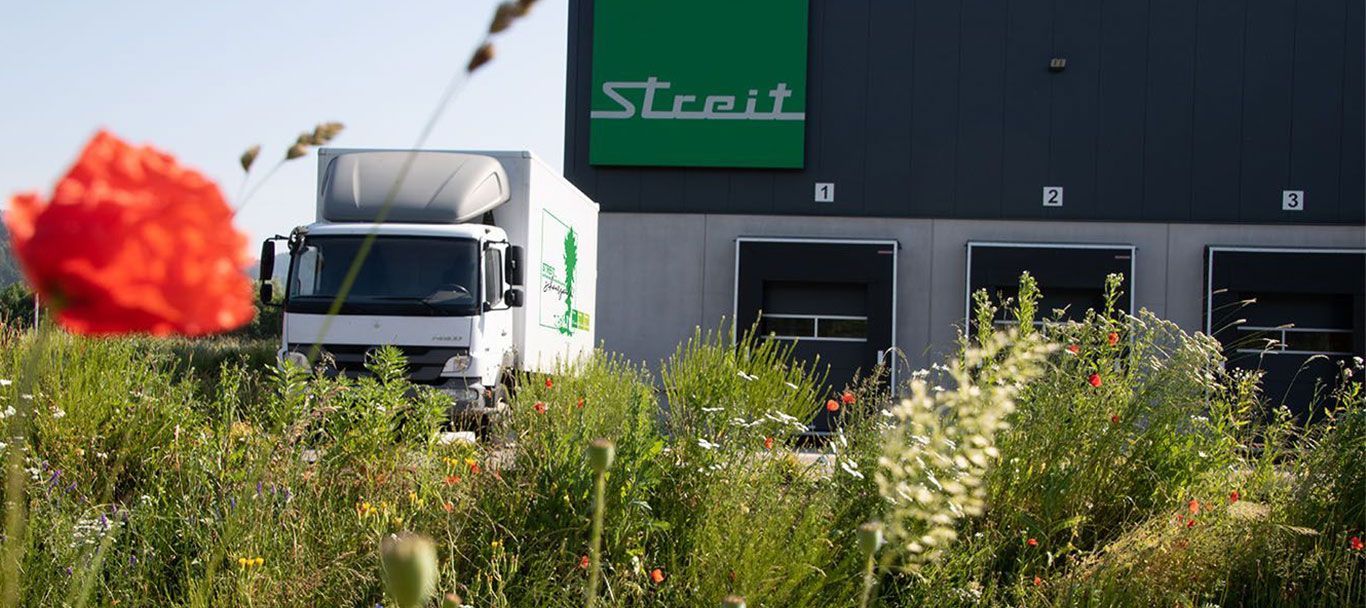 Wiese mit Blumen, dahinter ein weißer LKW und ein Gebäude mit dem Logo der Firma Streit Service & Solution
